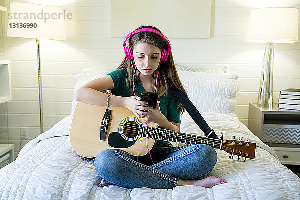 Teenager-Mädchen benutzt Smartphone  während sie mit Gitarre im Schlafzimmer sitzt