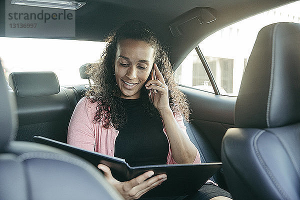 Geschäftsfrau sieht sich Datei an  während sie im Auto mit einem Smartphone telefoniert