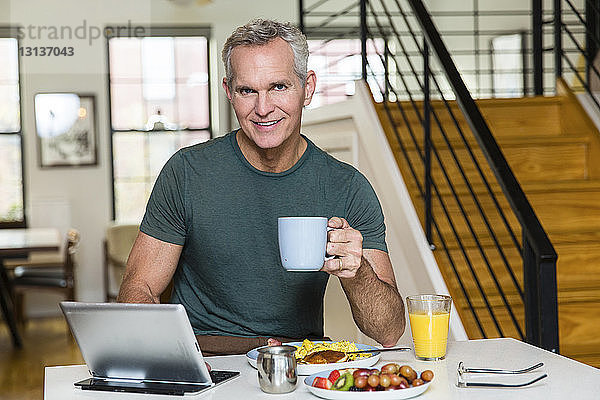 Porträt eines reifen Mannes  der eine Kaffeetasse hält  während er einen Tablet-Computer auf dem Esstisch zu Hause benutzt