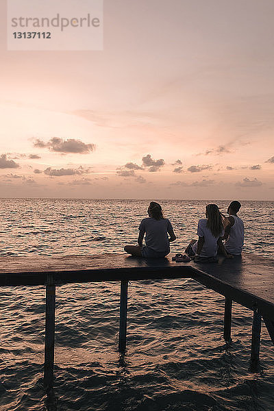 Scherenschnitt-Freunde sitzen bei Sonnenuntergang am Pier über dem Meer gegen den Himmel