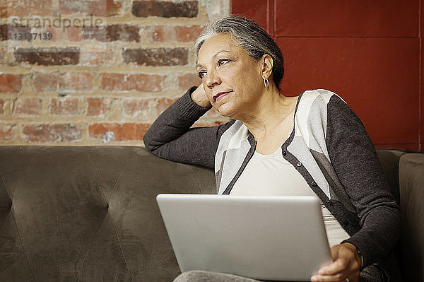 Frau mit Laptop-Computer schaut weg  während sie im Büro auf dem Sofa sitzt
