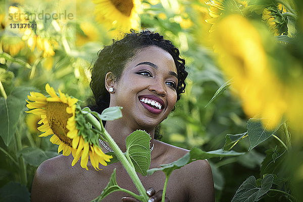 Nahaufnahme einer glücklichen Braut  die wegschaut  während sie inmitten von Sonnenblumen auf einem Bauernhof steht