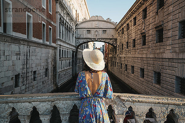 Rückansicht einer Frau mit Hut  die an einem Geländer auf einer Brücke inmitten von Gebäuden steht