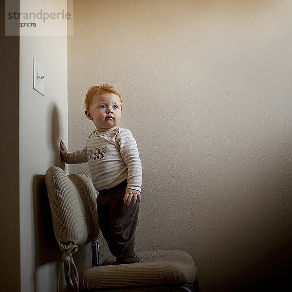 Süßer kleiner Junge schaut weg  während er zu Hause auf einem Stuhl an der Wand steht