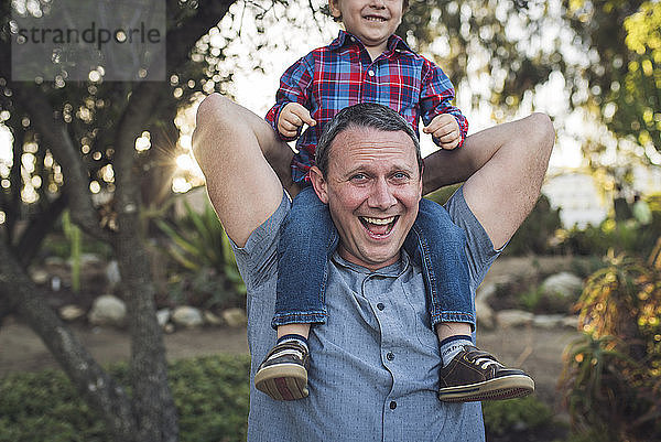 Porträt eines fröhlichen Vaters  der seinen Sohn auf den Schultern trägt  während er mit ihm im Park spielt