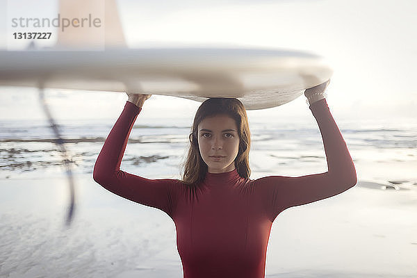 Porträt einer jungen Frau  die am Strand stehend ein Surfbrett auf dem Kopf trägt