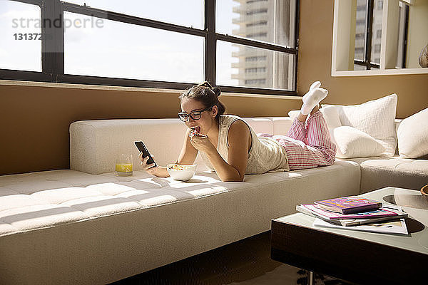 Frau benutzt Smartphone  während sie zu Hause auf dem Sofa frühstückt