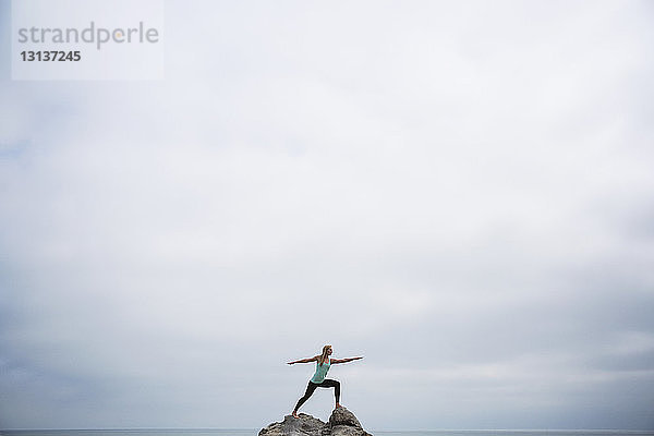 Frau übt die Pose von Worrier 2 auf Felsen im Meer vor bewölktem Himmel