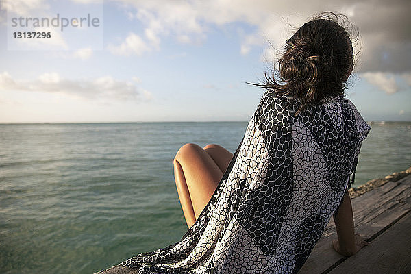 Rückansicht einer in eine Decke gehüllten Frau  die sich auf einer Strandpromenade am Meer entspannt