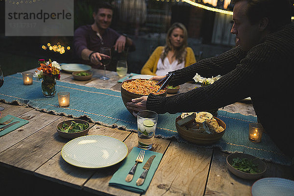 Mann hält Schüssel auf dem Tisch  während er mit Freunden auf der Terrasse zu Abend isst