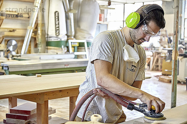 Schreiner mit Ohren- und Augenschutz beim Schleifen von Holzbrettern in der Werkstatt