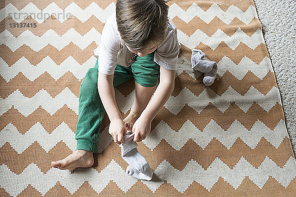 Schrägansicht eines Jungen  der eine Socke trägt  während er zu Hause auf dem Teppich sitzt