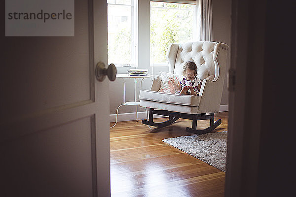 Mädchen liest Bilderbuch  während sie zu Hause auf einem Schaukelstuhl sitzt und durch die Tür gesehen wird
