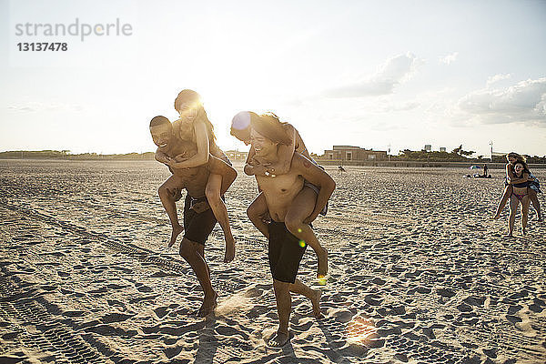 Freunde genießen Huckepack-Fahrten am Strand gegen den Himmel an sonnigen Tagen