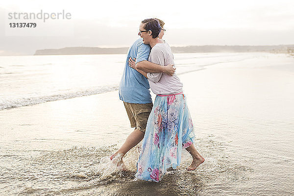 Seitenansicht eines glücklichen Paares  das sich umarmt  während es am Strand am Ufer steht