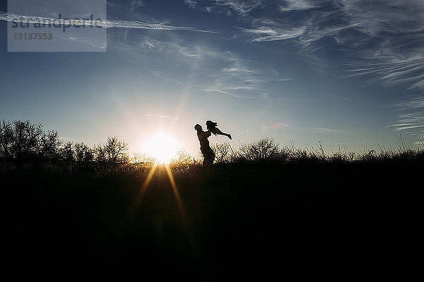 Silhouettenvater hebt Tochter hoch  während er mit ihr bei Sonnenuntergang auf dem Feld gegen den Himmel spielt