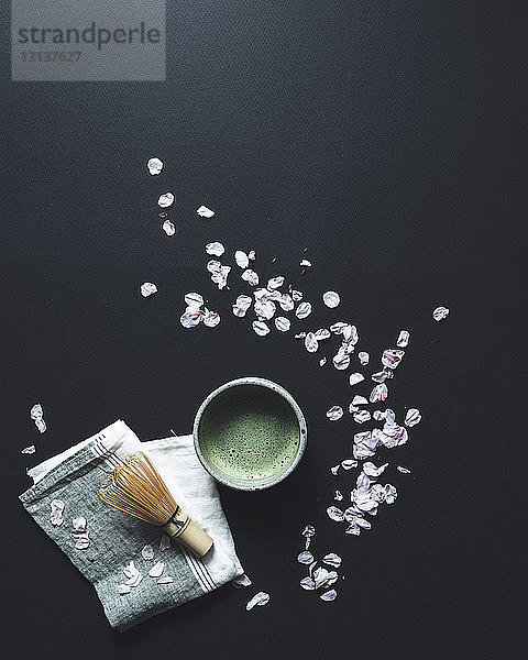 Draufsicht auf Matcha-Tee und Bambusbesen durch Blütenblätter auf schwarzem Hintergrund