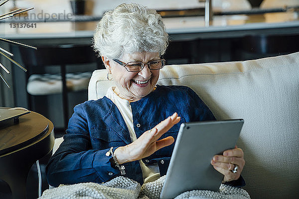 Glückliche ältere Frau bei Videokonferenzen am Tablet-Computer  während sie sich zu Hause auf dem Sofa entspannt