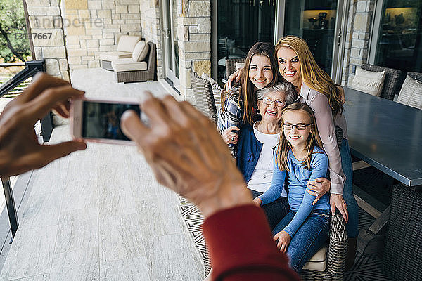 Abgetrennte Hände eines älteren Mannes fotografieren glückliche Familie durch ein Smartphone auf der Veranda