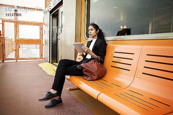 Betrachtete Frau benutzt digitales Tablett  während sie auf einer orangefarbenen Bank in der Stadt sitzt