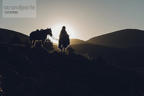 Scherenschnittfrau mit Pferd steht bei Sonnenuntergang auf Berg vor klarem Himmel