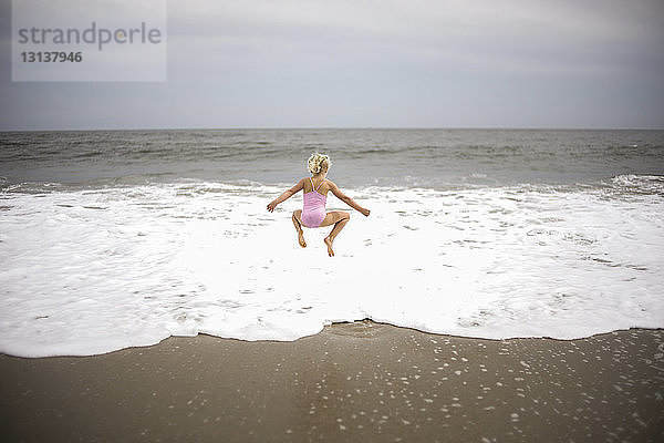 Rückansicht eines verspielten Mädchens  das am Strand am Ufer gegen den Himmel springt
