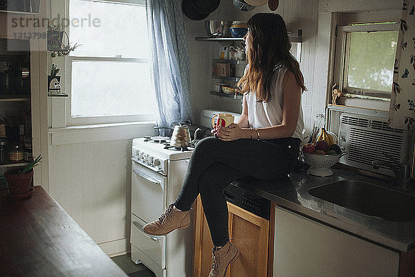 Nachdenkliche Frau hält Kaffeetasse  während sie auf der Küchentheke sitzt