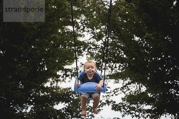 Porträt eines süßen kleinen Jungen  der sich auf dem Spielplatz gegen Bäume schwingt
