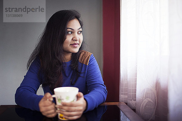 Lächelnde  nachdenkliche Frau hält Kaffeetasse  während sie am Tisch sitzt