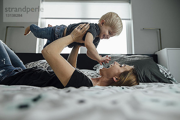 Seitenansicht einer glücklichen Mutter  die ihren Sohn abholt  während sie zu Hause im Bett liegt