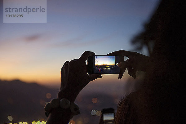 Frau fotografiert beleuchtete Stadtlandschaft mit Smartphone bei Sonnenuntergang