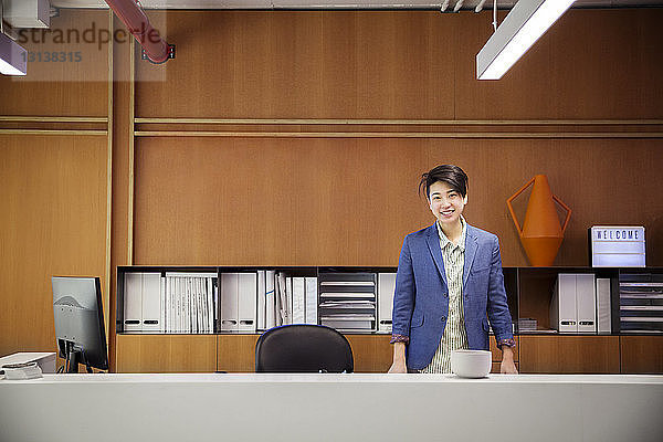 Porträt einer selbstbewusst lächelnden Geschäftsfrau  die im Büro am Schreibtisch steht