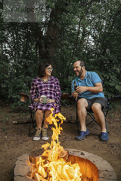 Das glückliche Paar sitzt in voller Länge auf Campingstühlen am Lagerfeuer im Wald