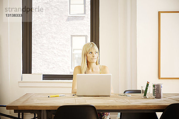 Nachdenkliche Geschäftsfrau sitzt mit Laptop am Schreibtisch im Kreativbüro