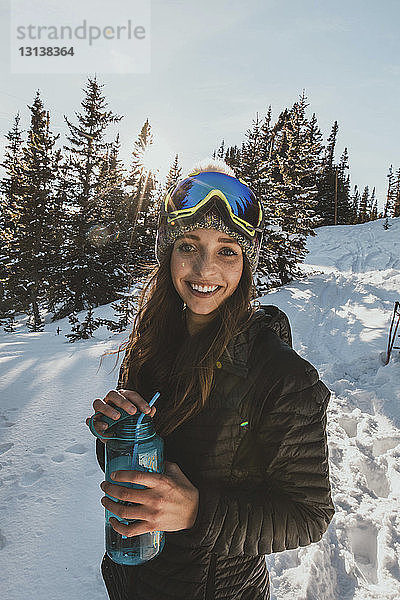 Porträt einer lächelnden jungen Frau mit Skibrille  die eine Wasserflasche auf einem verschneiten Feld hält