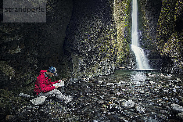 Mann liest Karte  während er am Wasserfall im Wald sitzt