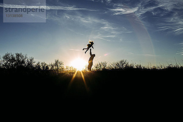 Verspielter Silhouettenvater  der seine Tochter in die Luft wirft  während er mit ihr bei Sonnenuntergang auf dem Feld gegen den Himmel spielt