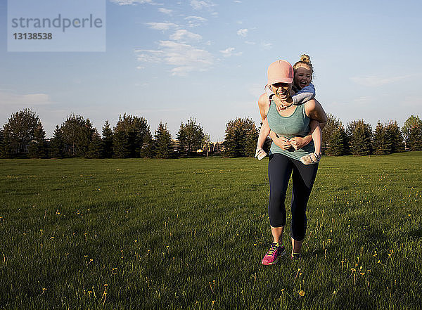 Glückliche Mutter nimmt ihre Tochter huckepack  während sie im Park auf einem Grasfeld gegen den Himmel trainiert