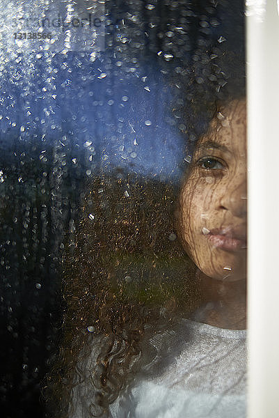 Mädchen schaut weg  während sie zu Hause steht und durch nasses Fensterglas gesehen wird