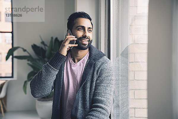 Lächelnder Geschäftsmann spricht am Smartphone  während er im Büro auf dem Fensterbrett sitzt