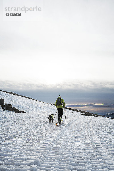 Mann wandert mit Hund auf schneebedecktem Feld vor bewölktem Himmel