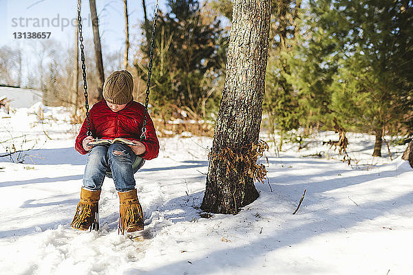 Mädchen liest ein Buch in voller Länge  während sie im Winter im Park auf der Schaukel sitzt