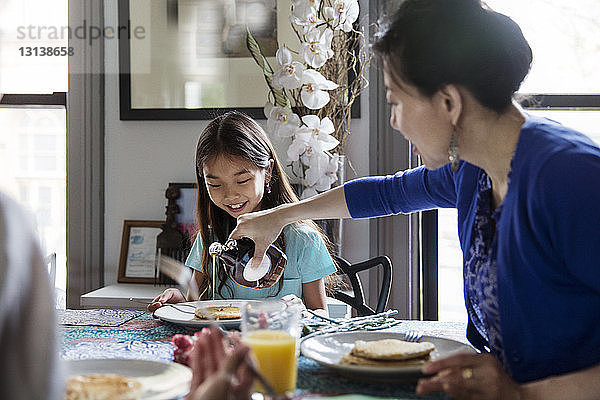 Mutter gießt beim Frühstück Ahornsirup auf Pfannkuchen für die Tochter