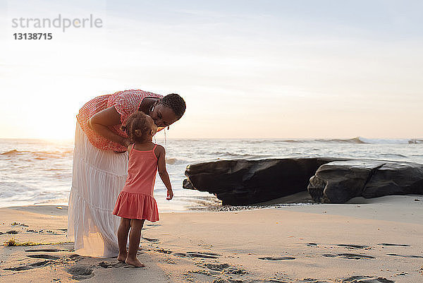 Mutter küsst Tochter  während sie am Strand gegen den Himmel steht