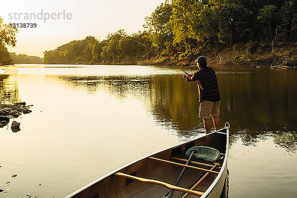 Rückansicht eines mittleren erwachsenen Mannes beim Angeln  während er bei Sonnenuntergang am Boot im See steht