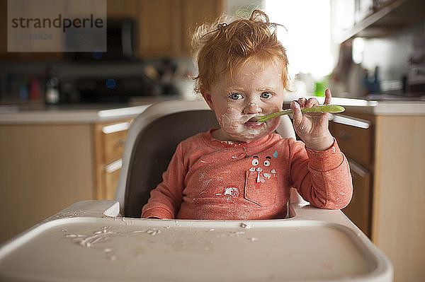 Porträt eines süßen kleinen Jungen mit unordentlichem Gesicht  der zu Hause auf einem Hochstuhl sitzend Essen isst