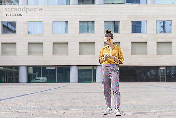 Selbstbewusste Geschäftsfrau benutzt Smartphone  während sie auf dem Bürgersteig gegen Gebäude in der Stadt steht