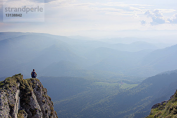 Rückansicht eines auf einer Klippe sitzenden Mannes vor bewölktem Himmel