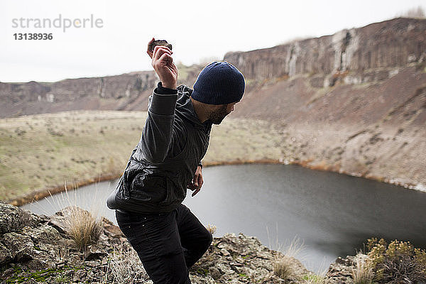 Seitenansicht eines Wanderers  der auf einer Klippe stehend einen Stein in den See wirft