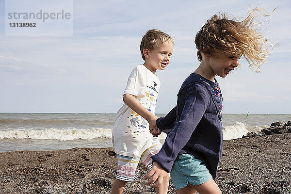 Glückliche Geschwister halten sich an sonnigen Tagen beim Spaziergang am Strand gegen den Himmel an den Händen